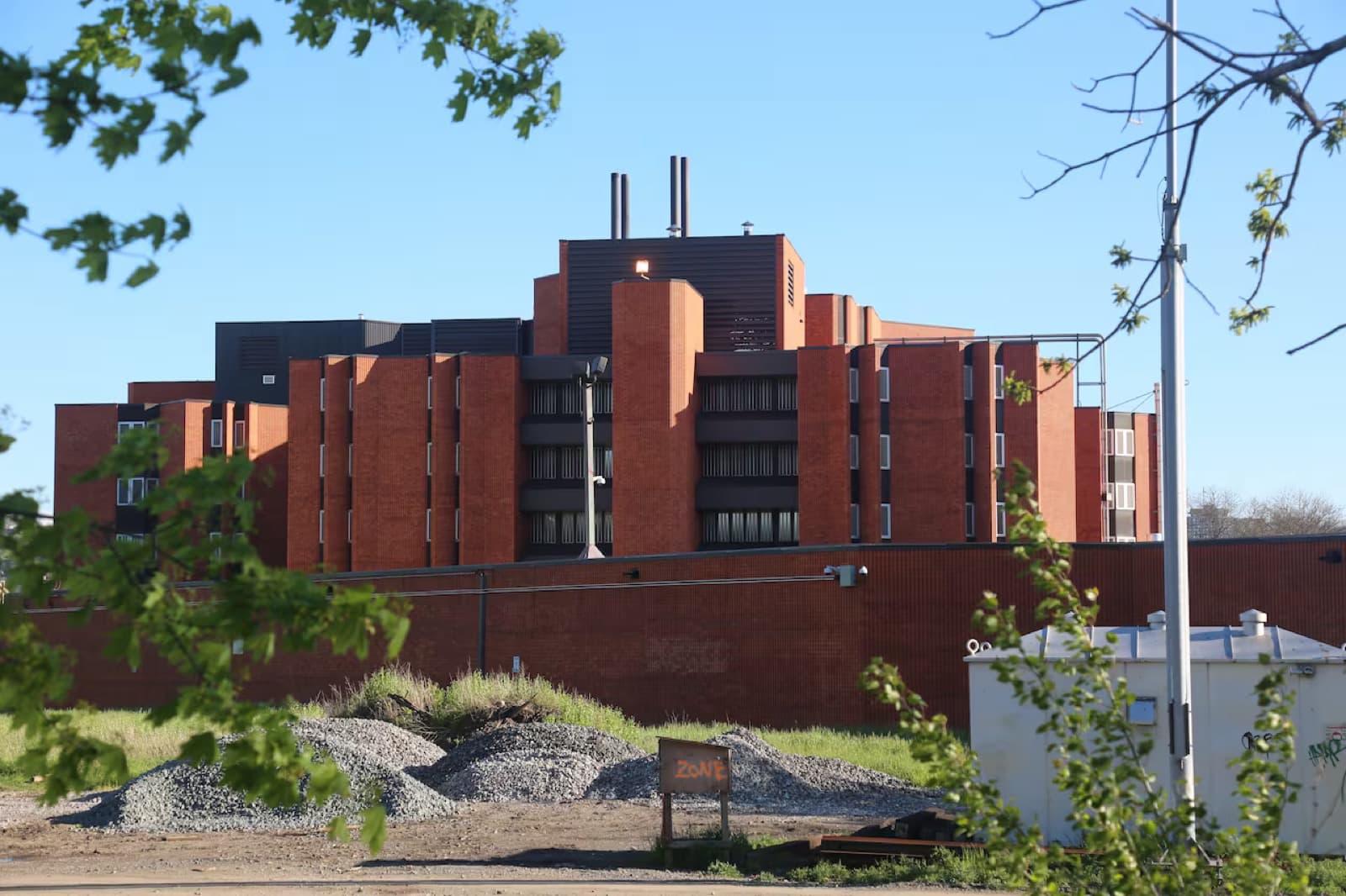 A large red-brick institutional building behind a perimeter wall, with gravel piles and a site trailer in front